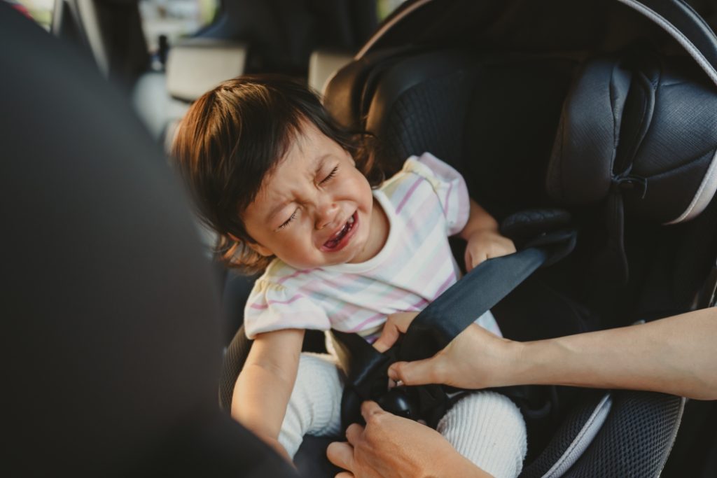 mother is fastening safety belt to crying toddler girl in a car seat, safety baby chair travelling