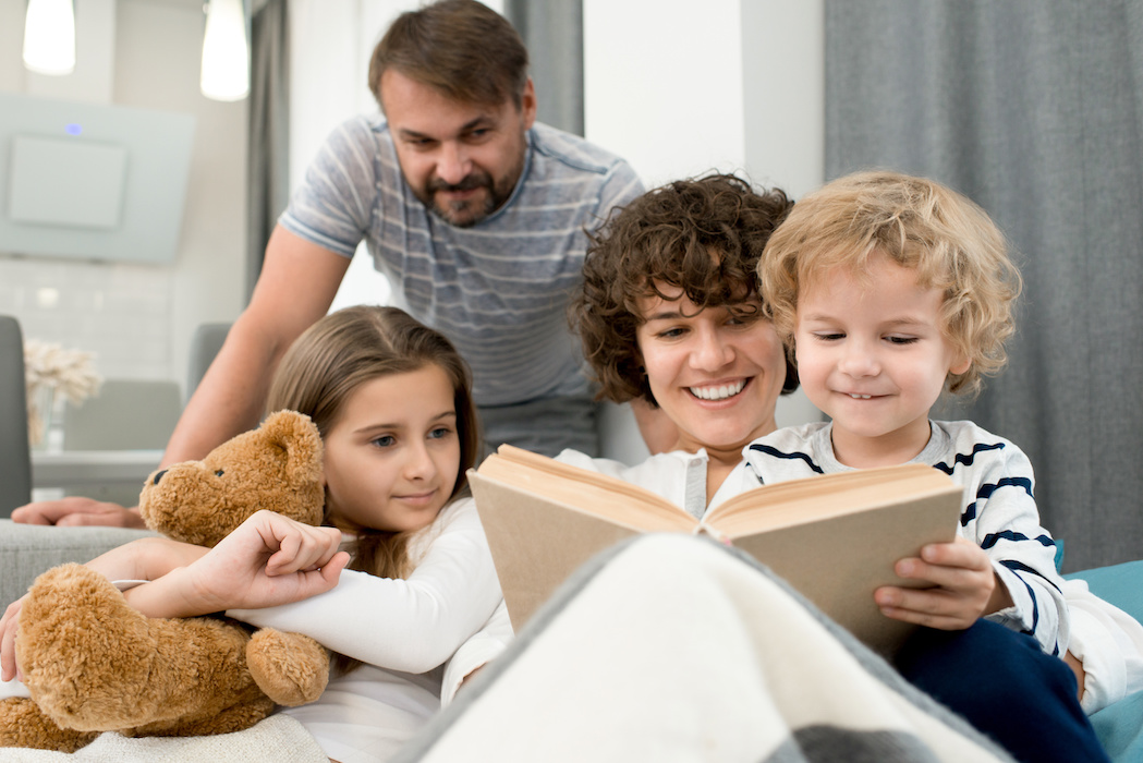 Loving family of four gathered together in cozy living room and reading adventure story aloud, pretty little girl embracing her teddy bear