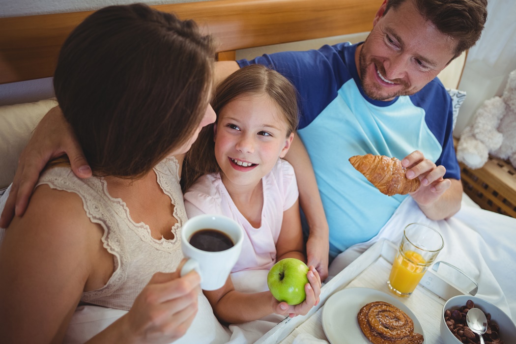 Parents sitting on bed with daughter and having breakfast