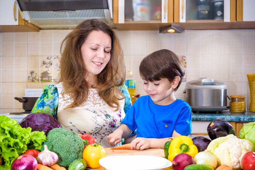 The young cook mother standing with her little son in the kitchen and salting vegetables