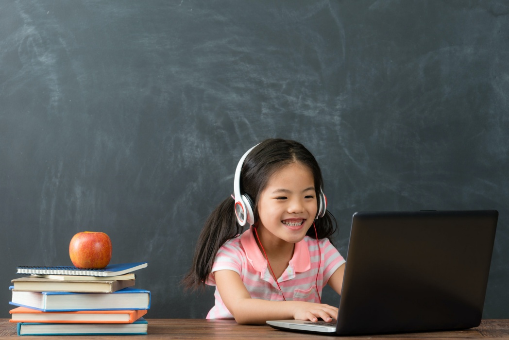 cheerful young little girl children using laptop computer with headphones studying through online e-learning system in chalkboard background.