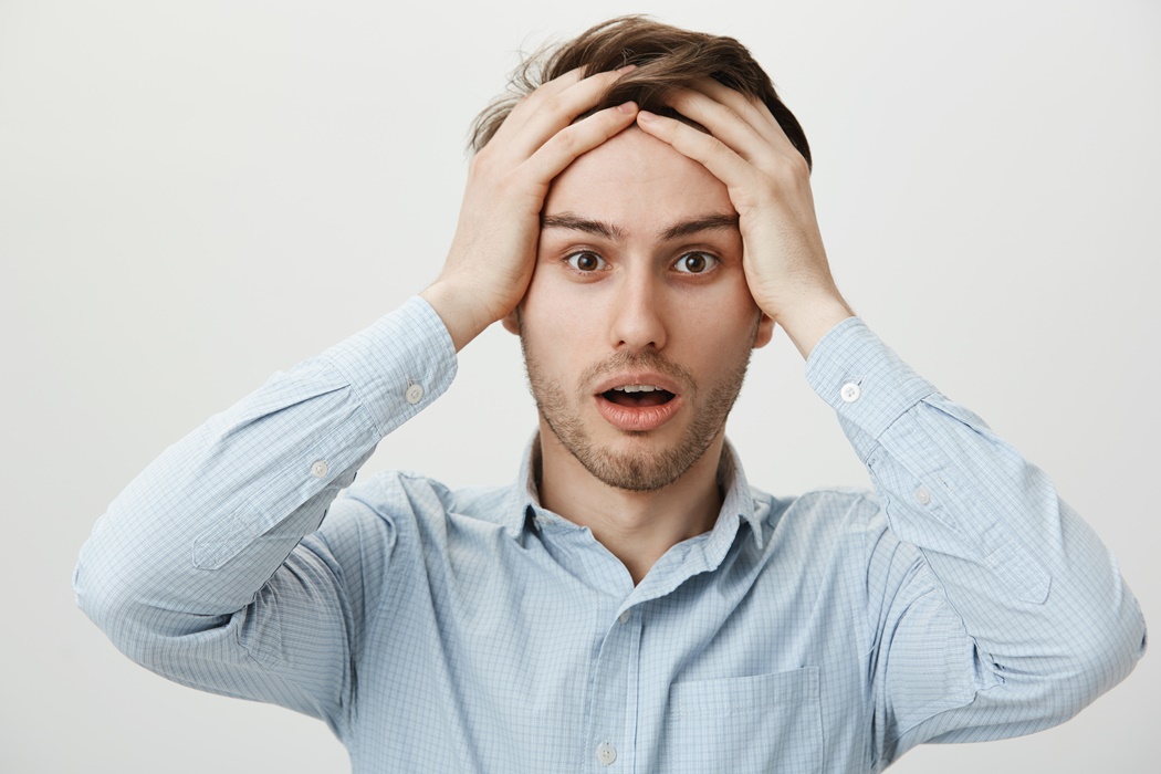 Despair and regret concept. Portrait of shocked attractive young businessman holding hands on forehead, looking with opened mouth, losing all investments and feel devastated, standing over gray wall