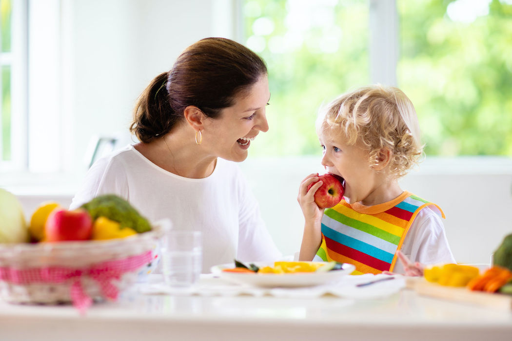 Mother feeding child vegetables. Mom feeds kid in white kitchen with window. Baby boy sitting in high chair eating healthy lunch of steamed carrot and broccoli. Nutrition, vegetarian diet for toddler
