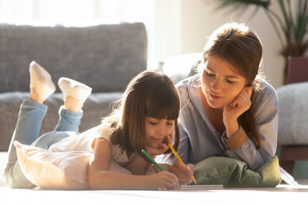 Mother or baby sitter teaching little girl laying on warm living room floor at home, focused mum helping kid daughter learning drawing with colored pencils together, mom and child creative activity