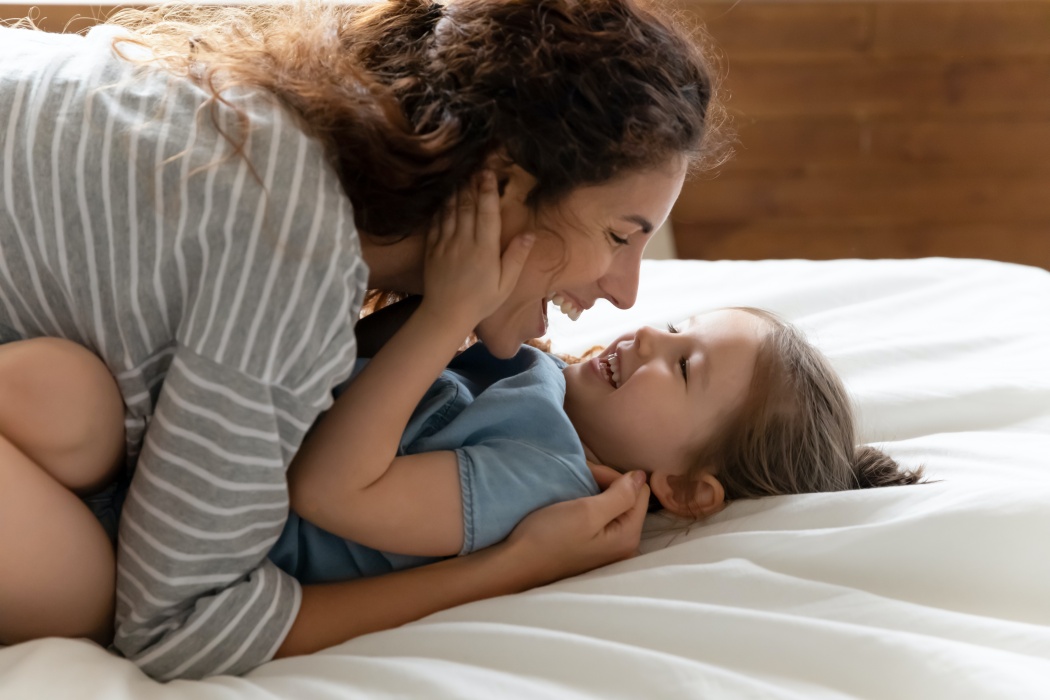 Head shot young happy mother having fun with joyful adorable little preschool child daughter on bed. Laughing attractive mommy cuddling small kid girl, playing spending free time together in bedroom.