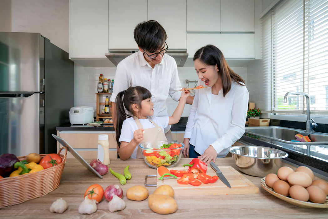 Asian daughters feeding salad to her mother and her father stand by when a family cooking in the kitchen at home. Family life love relationship, or home fun leisure activity concept