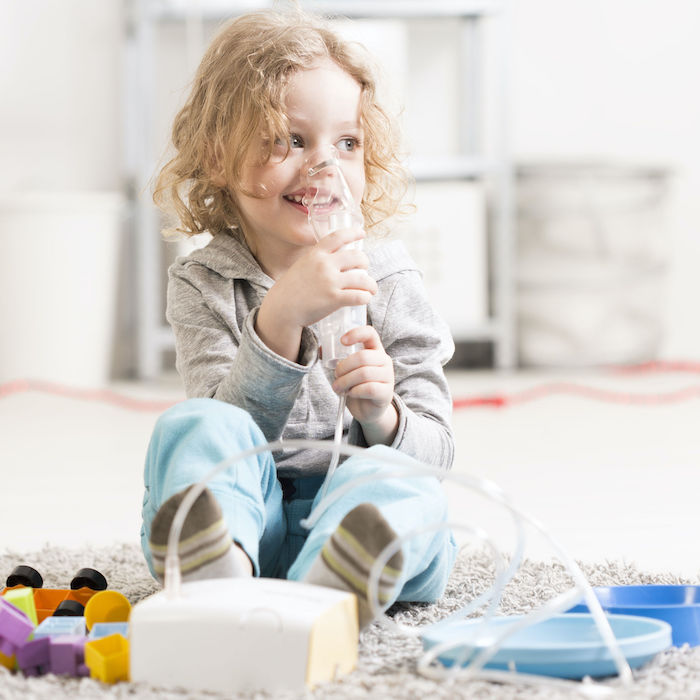 Small girl doing inhalation, sitting on a floor among toys in light interior