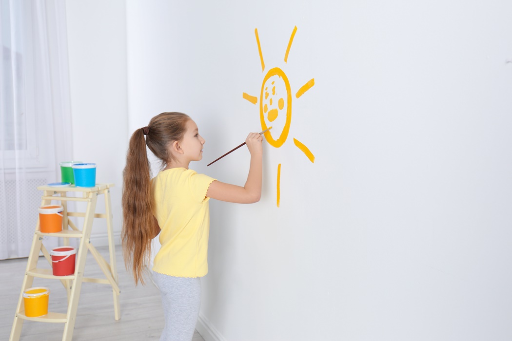 Little child painting sun on white wall indoors