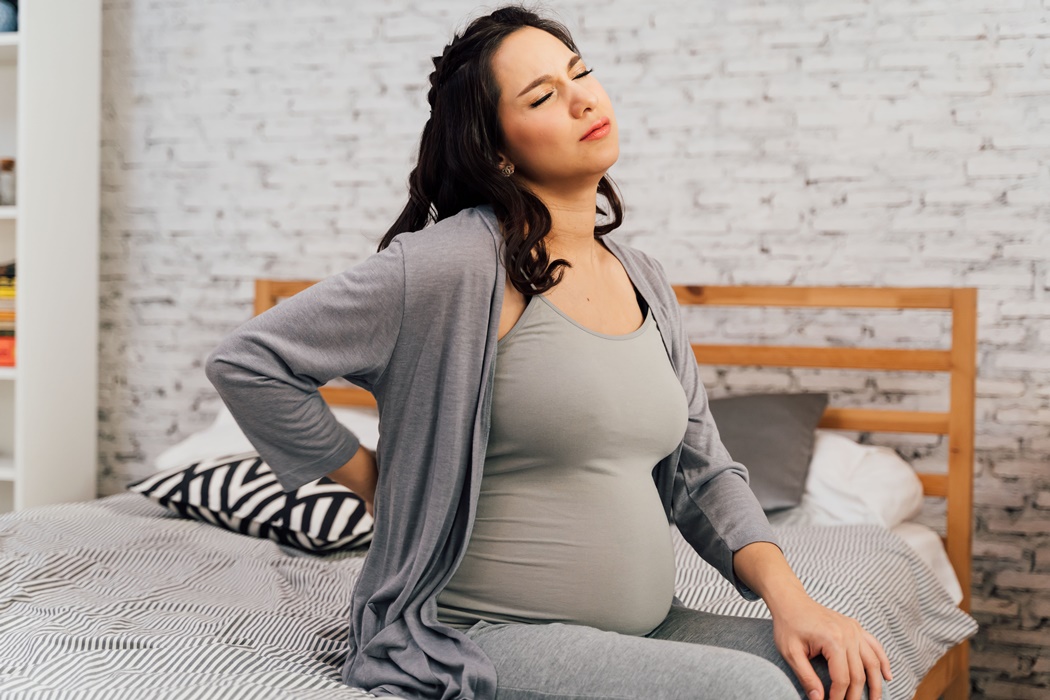 Asian mixed Caucasian pregnant woman suffering back pain sits on bed at home.