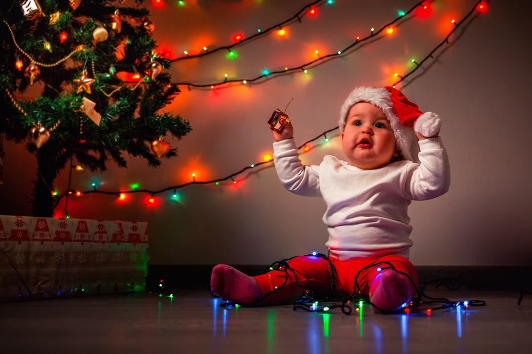 Christmas. A pretty baby in a Christmas hat sits near a Christmas tree decorated with lighting garland. The concept of winter Christmas holidays
