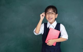 Asian Chinese little Girl in uniform standing against green blackboard in classroom