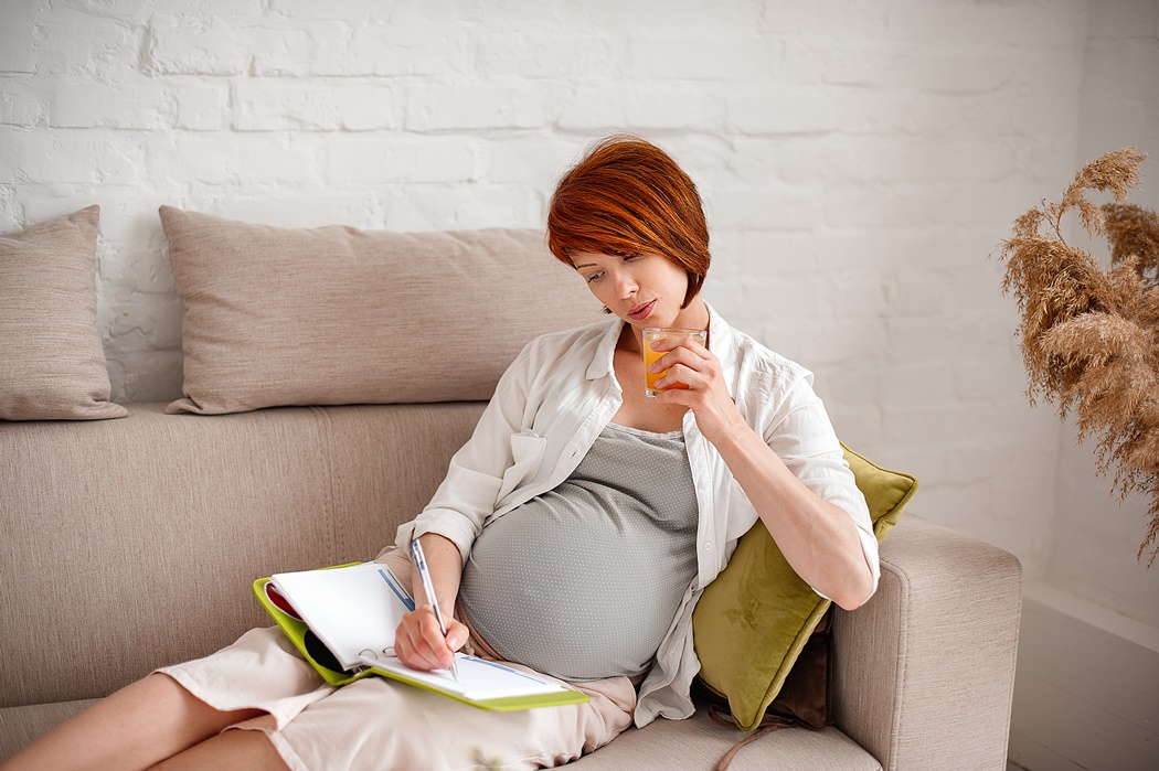Pregnant woman drinks juice and writes plans in a diary while lying on a sofa