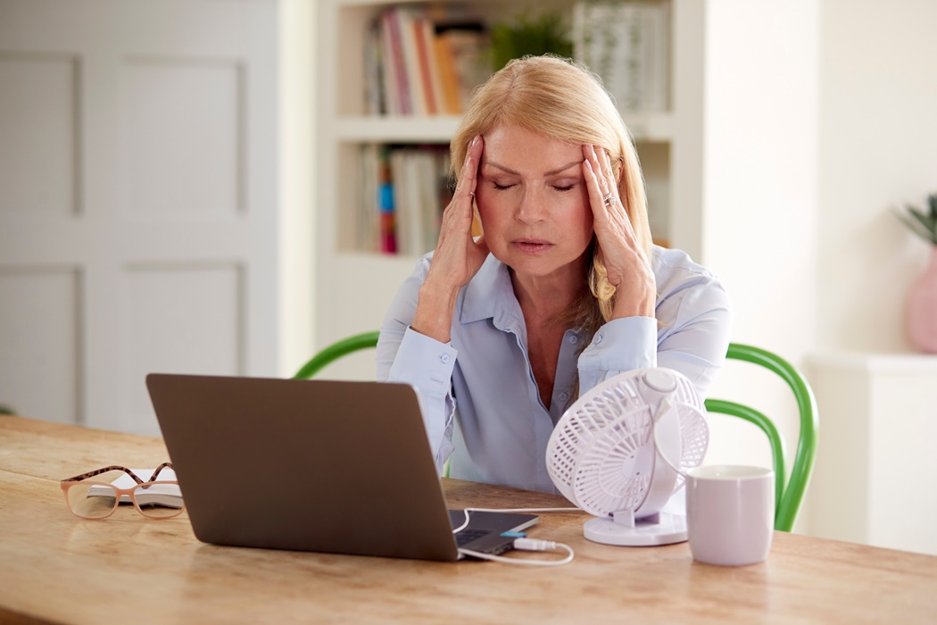 Menopausal Mature Woman Having Hot Flush At Home Cooling Herself With Fan Connected To Laptop