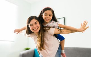 Carefree mom and little girl laughing while playing at home. Hispanic beautiful mother carrying her daughter on the back like an airplane